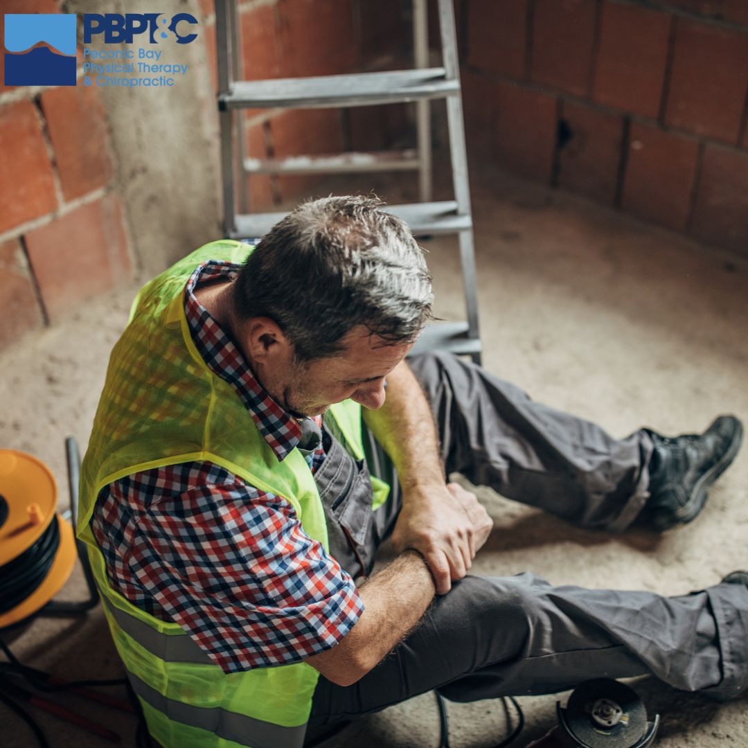 A construction worker in a safety vest sits on the floor holding his knee in pain, with a ladder and tools nearby—he may need to see a Workers Compensation Doctor in Holbrook. A construction worker in a safety vest sits on the floor holding his knee in pain, with a ladder and tools nearby—he may need to see a Workers Compensation Doctor in Holbrook