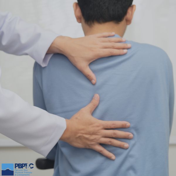 A healthcare professional examines a patient’s upper back by placing hands on the patient's back while the patient sits facing away.