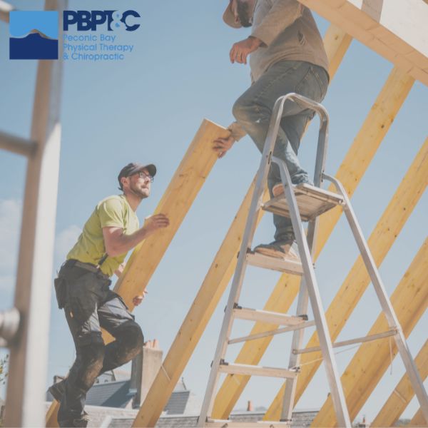 Two construction workers position a wooden beam on a roof frame; one stands on a ladder while the other supports the beam from the ground. The PBPT&C logo appears in the top left corner. Workers Compensation Doctor in Patchogue
